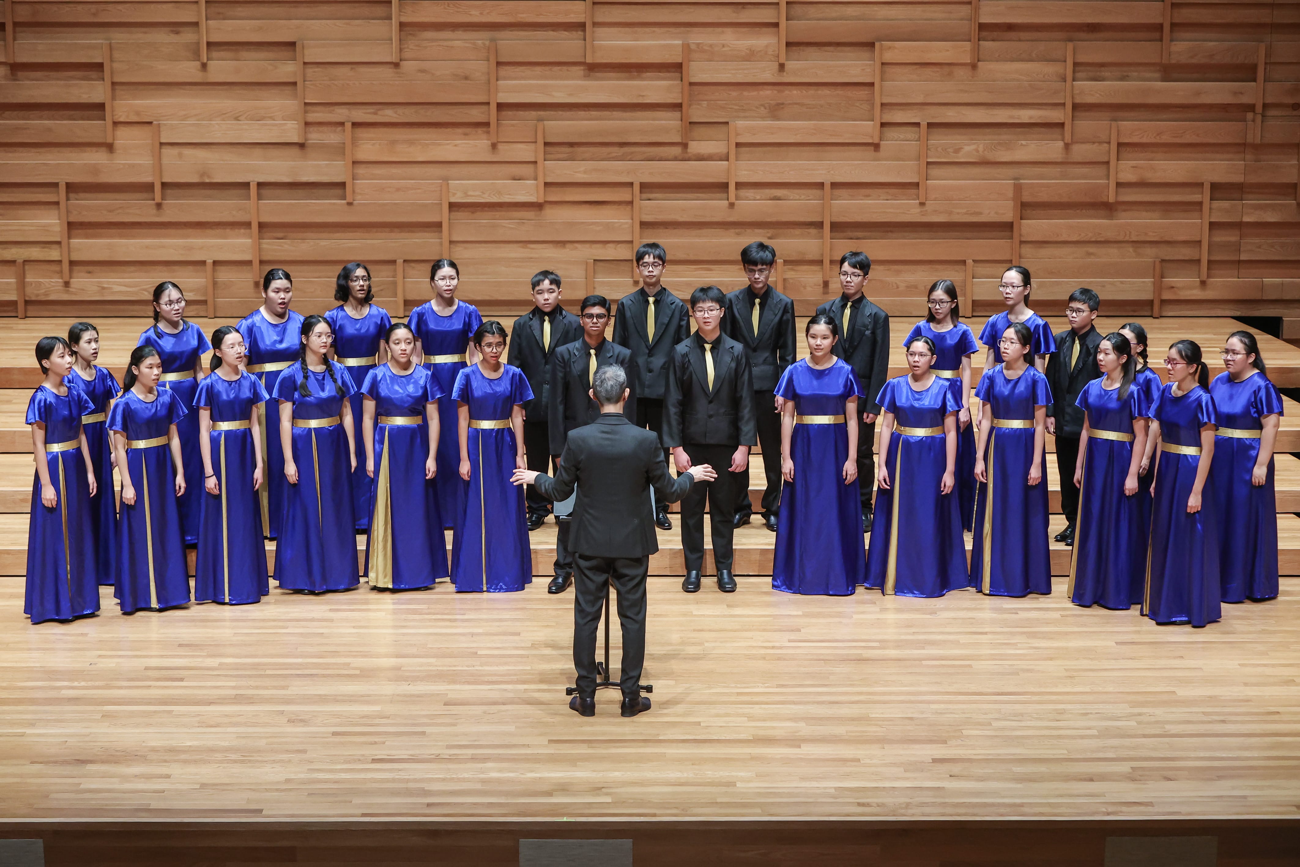 A conductor leads a youth choir on a wooden stage; girls wear blue and gold gowns, boys wear black suits with gold ties.